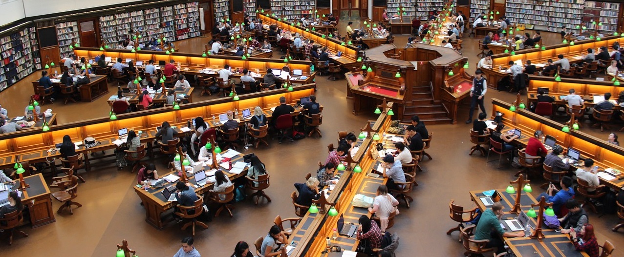 Biblioteca universitaria con mesas distribuidas formando una estrella. Hay muchos estudiantes
